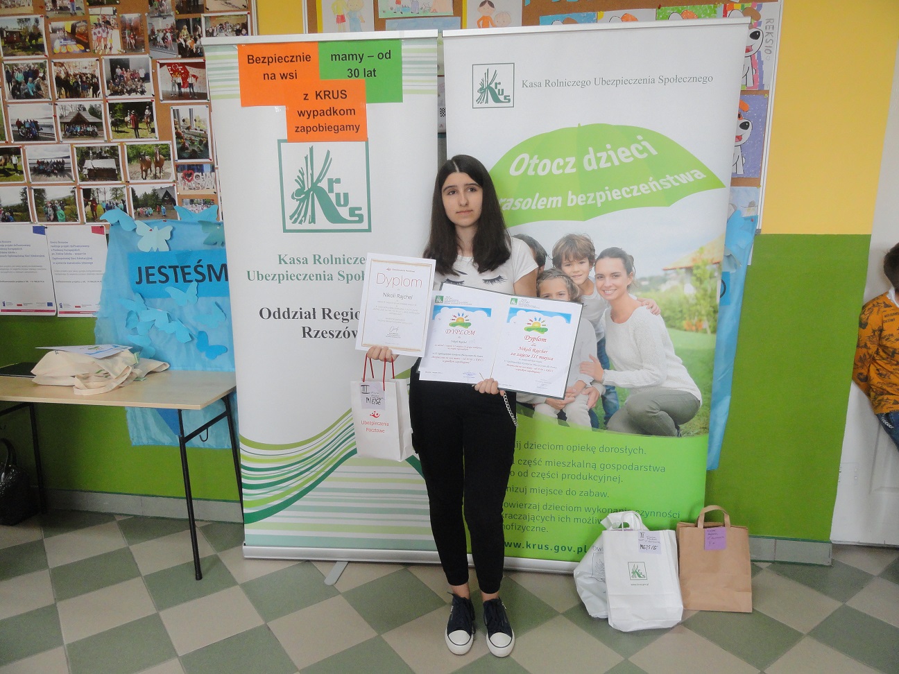 Dziewczyna w czarnych spodniach i białej bluzce stoi przed tablicami promującymi KRUS.