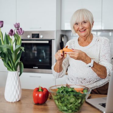 Starsza kobieta siedząca przy stole w kuchni. Na stole laptop, kwiaty i warzywa.