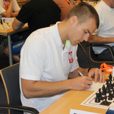 Młody mężczyzna grający w szachy. W tle inne grajace osoby.