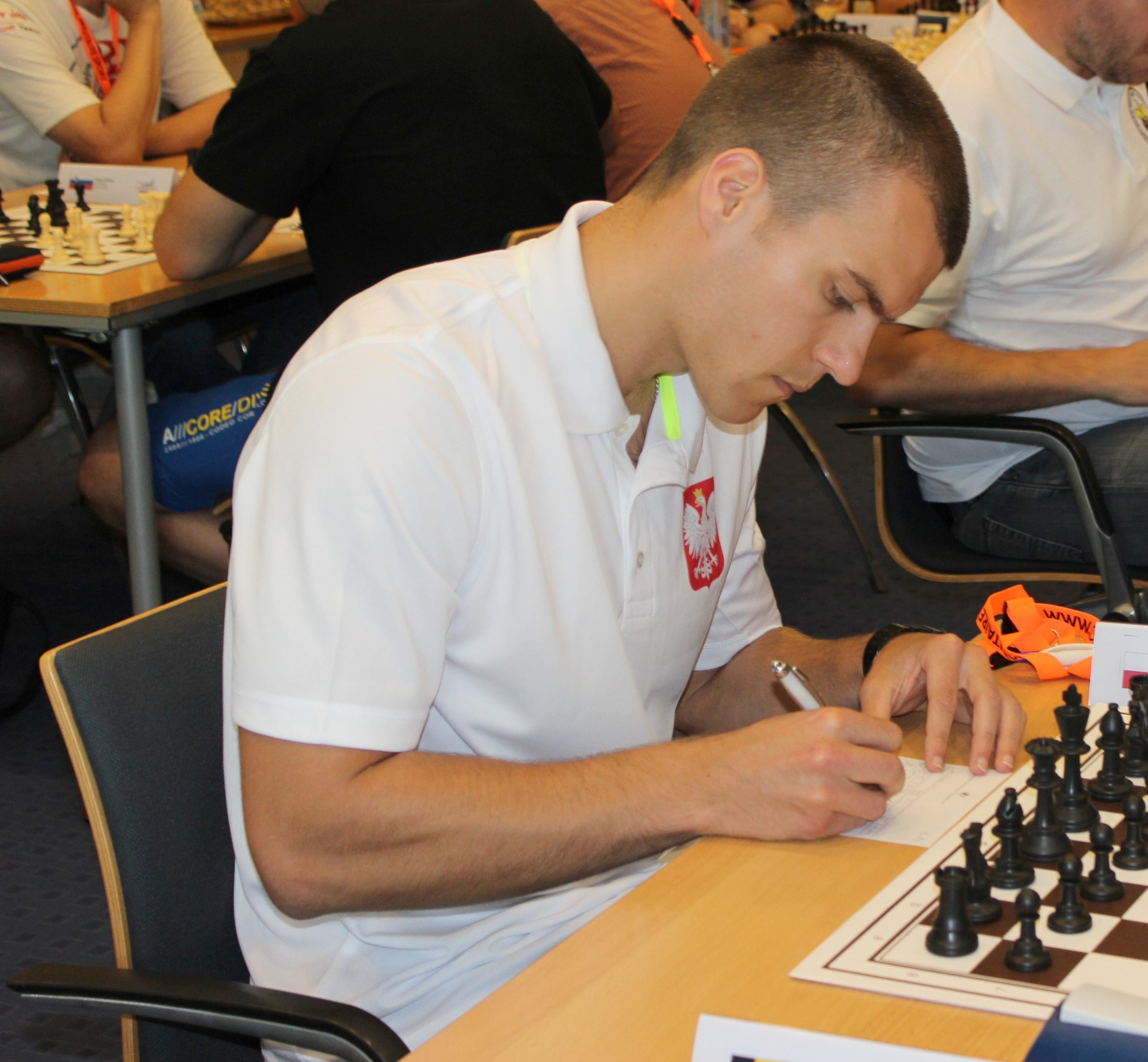 Młody mężczyzna grający w szachy. W tle inne grajace osoby.