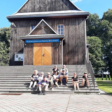 Grupa osób siedzi na schodach do drewnianego kościoła. Obok niego rosną drzewa liściaste.