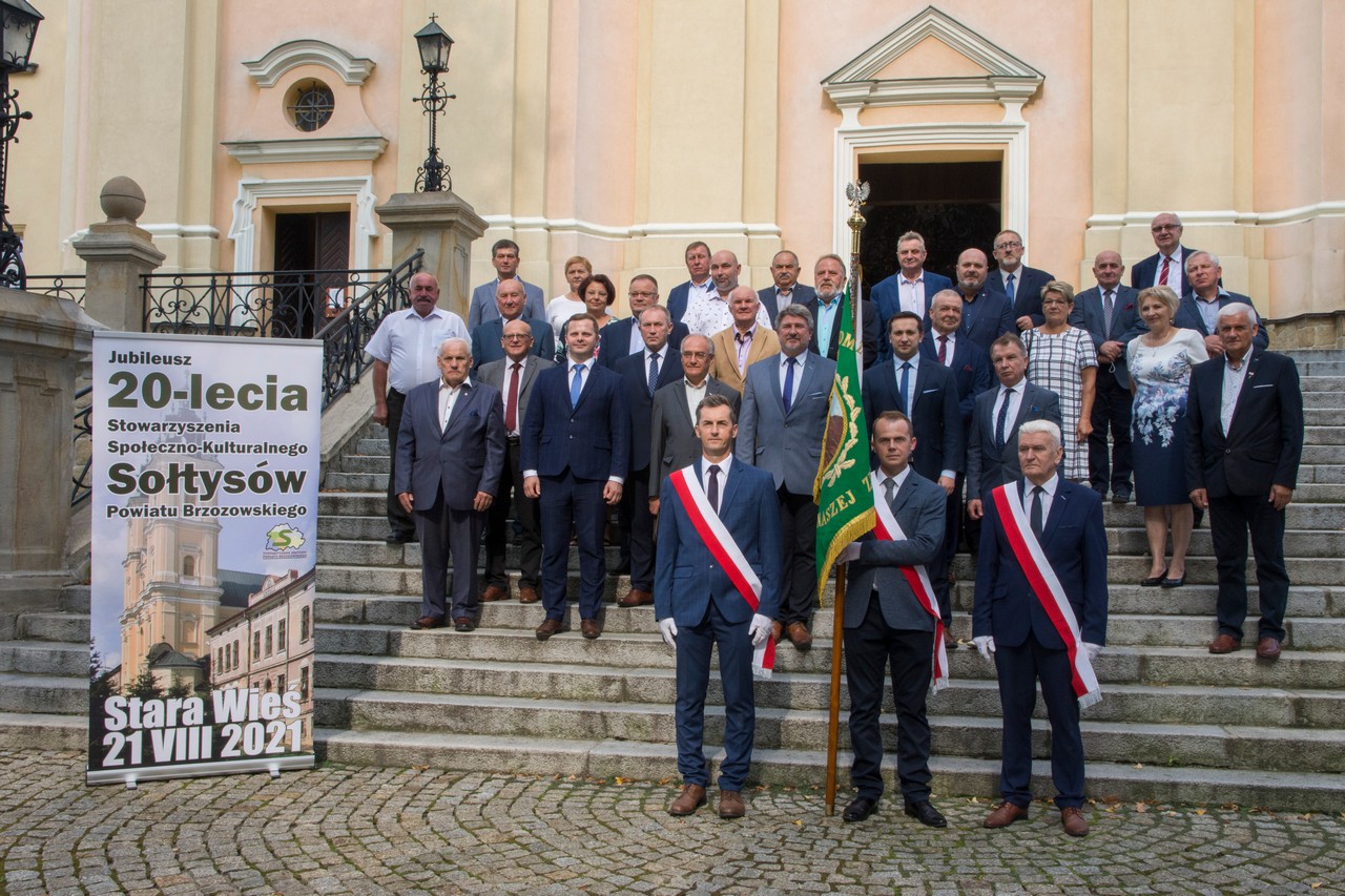 Grupa osób odświętnie ubranych stoi na schodach przed ogromnym kościołem z jasną elewacją. Przed nimi stoją trzy osoby, które trzymają sztandar.