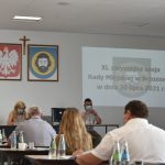 Grupa siedzących dorosłych osób. W tle herby i promień rzutnika.