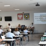 Grupa siedzących dorosłych osób. W tle herby i promień rzutnika.