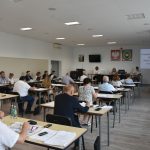 Grupa siedzących osób, odświętnie ubranych. Jeden ze stojacych mężczyzn trzyma mikrofon.