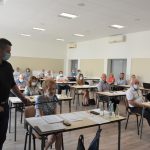 Grupa siedzących osób, odświętnie ubranych. Jeden ze stojacych mężczyzn trzyma mikrofon.