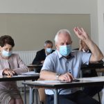 Starszy mężczyzna w maseczce siedzi przy stoliku w jasnym pomieszczeniu na sesji, za nim kobieta w maseczce. Mężczyzna podnosi rękę do góry.