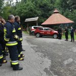 Grupa osób w strojach strażackich stoi na odprawie w lesie. Przed nimi stoi 5 mężczyzn, którzy podsumowują działania. Z lewej strony widac samochody strażackie. Za nimi drzewa liściaste.