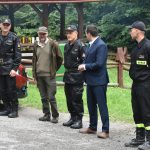 Grupa mężczyzn stoi na leśnej polanie. Obok nich zaparkowany jest samochód pożarniczy, za nimi rosną drzewa.