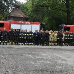 Grupa kilkudziesięciu strażaków i gosi pozuje do zdjęcia na tle samochodów pożarniczych.