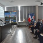 Grupa osób siedzi na krzesłach w jasnej sali i patrzy na prezentację multimedialną wyświetlaną na ścianie.