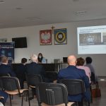 Grupa osób siedzi w jasnej sali i patrzy na prezentację multimedialną.