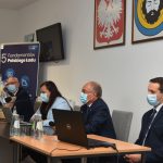 Grupa osób siedzi przy stoliku, za nimi na ścianie wiszą obrazy godła i herbu gminy.