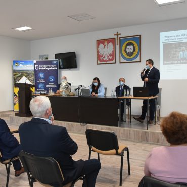 Grupa osób siedzi w jasnej sali, przed nimi na prezydium siedzą trzy osoby i jeden młody mężczyzna przemawia do mikrofonu.