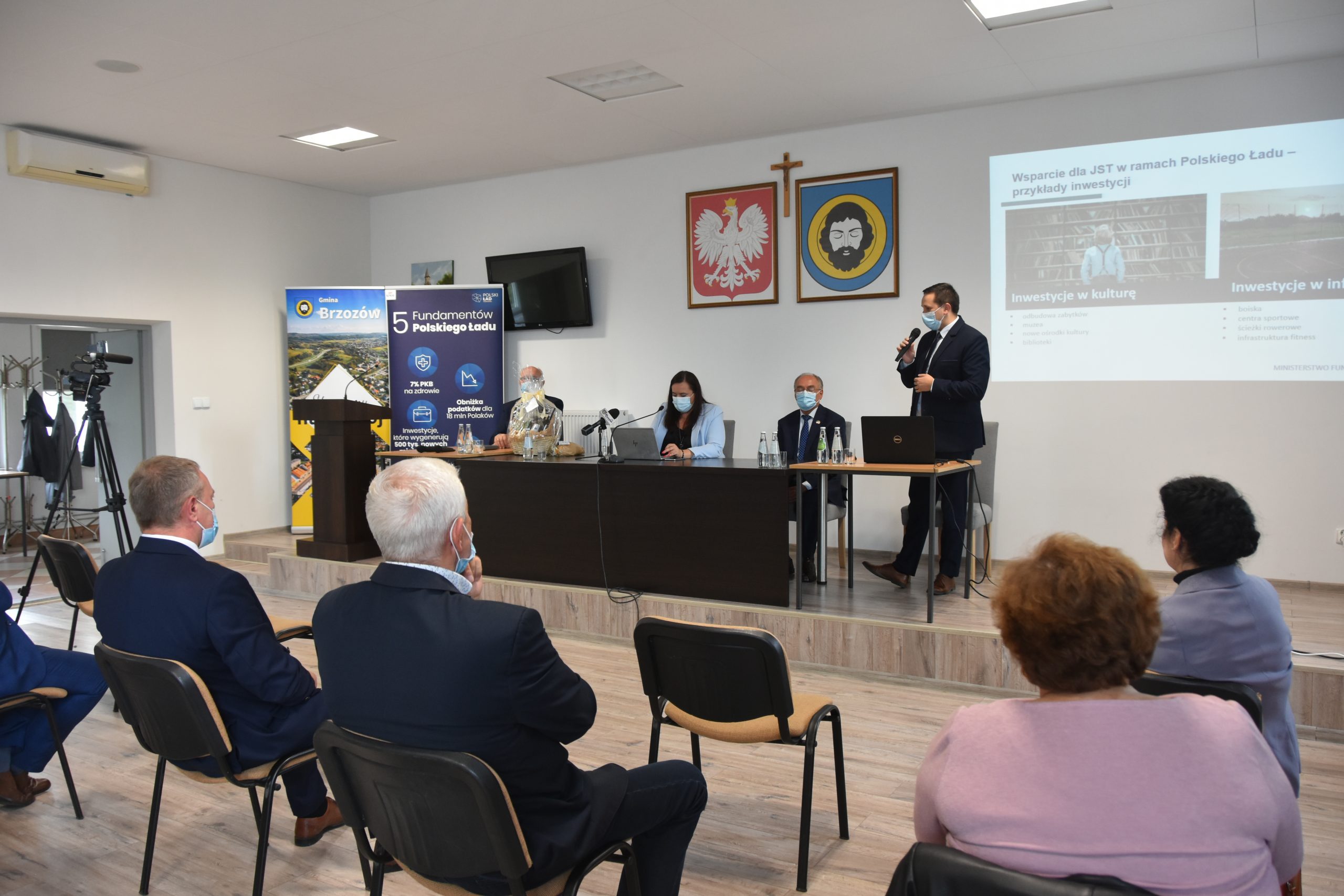 Grupa osób siedzi w jasnej sali, przed nimi na prezydium siedzą trzy osoby i jeden młody mężczyzna przemawia do mikrofonu.