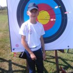 Chłopiec w stroju sportowym i czapce z daszkiem na głowie stoi na zewnątrz przed tarczą do strzelania z łuku.