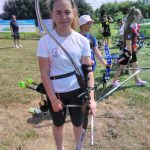 Dziewczynka w stroju sportowym, trzymająca w rękach łuk do strzelania stoi na trawiastym boisku. Za nią stoi grupa osób z łukami do strzelania.