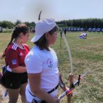 Dziewczynka w białym sportowym stroju i czapce z daszkiem stoi na trawiastym boisku i strzela z łuku.