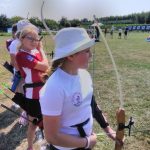 Dziewczynka w białym sportowym stroju i białym kapeluszu stoi na trawiastym boisku i strzela z łuku.