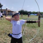 Chłopiec w białym stroju sportowym i kapeluszu stoi na trawiastym boisku i strzela z łuku.