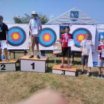 Grupa dzieci w strojach sportowych stoi na podium. Za nimi tarcze do strzelania z łuku.