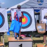 Chłopiec w stroju sportowym stoi na podium 1 miejsce. W rękach trzyma dyplom i nagrodę rzeczową. Za nimi tarcze do strzelania z łuku.