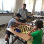 Chłopcy siedzą przy stoliku i grają w szachy. Przygląda im się straszy mężczyzna.