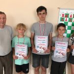 Grupa dzieci i starszy mężczyzna stoją przy ścianie i pozują do zdjęcia. Dzieci trzymają dyplomy i mają zawieszone na szyi medale.