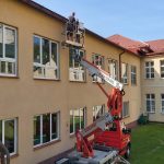 Mężczyźni na wyciągarce montują rynny przy dachu w szkole.