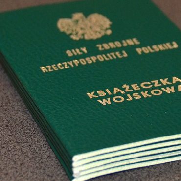 Książeczka wojskowa koloru zielonego leży na drewnianym blacie.