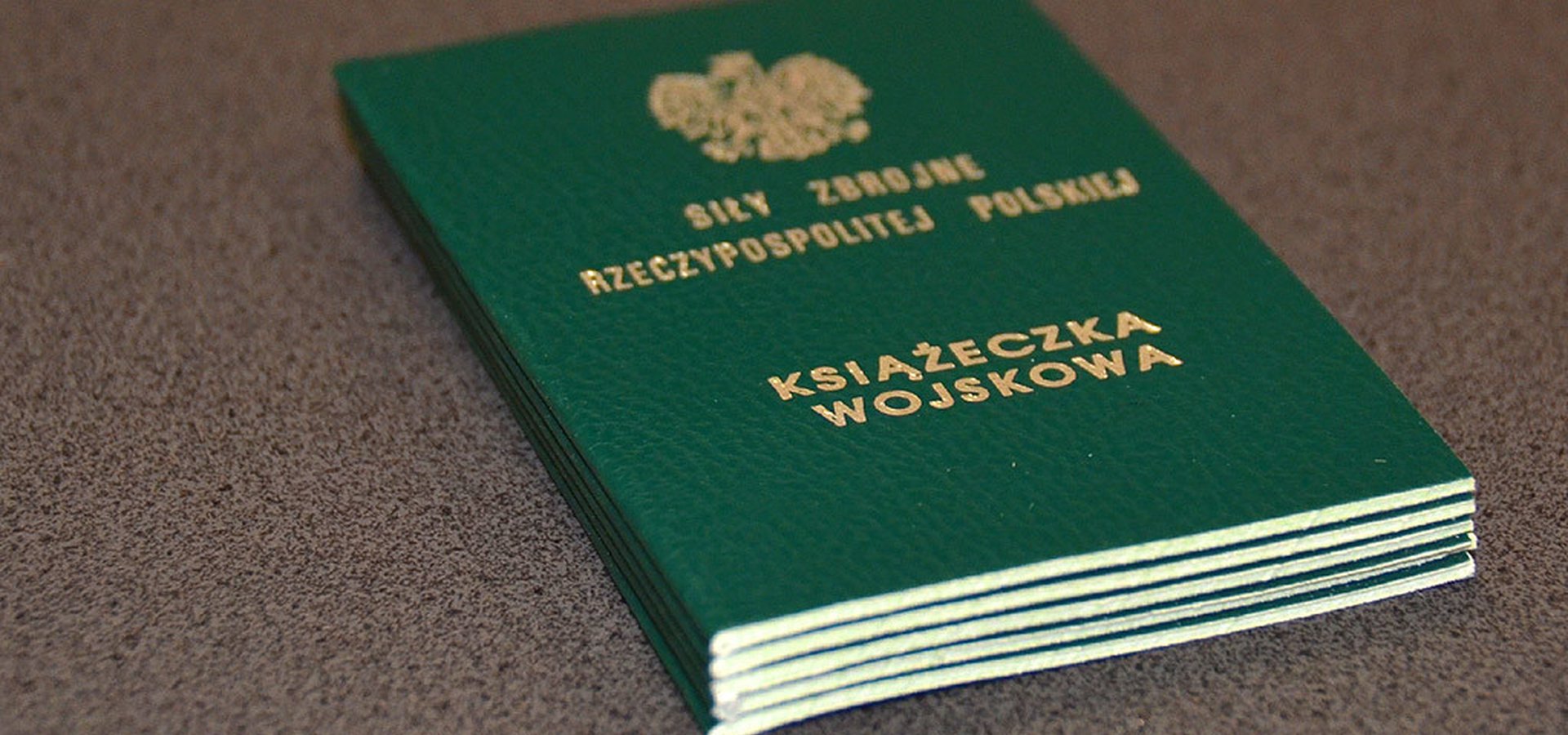 Książeczka wojskowa koloru zielonego leży na drewnianym blacie.