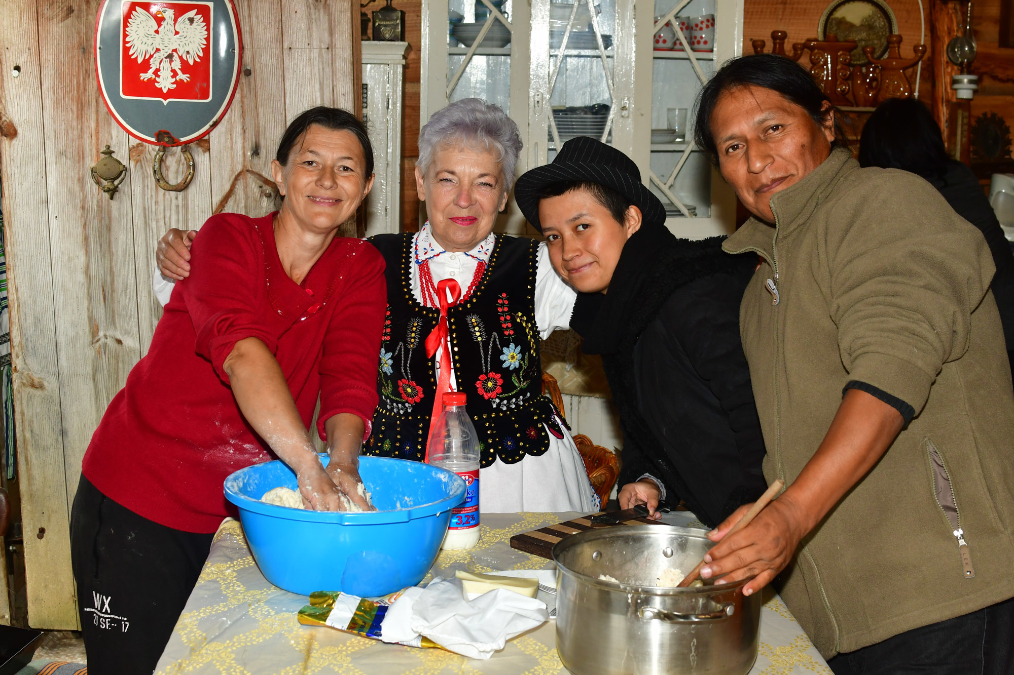 Grupa czterech osób stoi przy stole i wyrabia ciasto na chleb. Na stole leży niebieska miska, a w niej ciasto. Stoi też garnek.