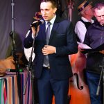 Młody mężczyzna w garniturze przemawia do mikrofonu. Za im stoi grupa osób w strojach ludowych.