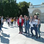 Sześcioosobowa grupa dzieci pozuje do zdjęcia na placu wyłożonym kamieniem, przed łukiem.