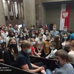 Grupa młodych ludzi siedzi w pomieszczeniu sejmu, za nimi wisi ogromna flaga z godłem Polski.