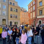 Grupa młodych ludzi podczas wycieczki stoi na placu z kostki brukowej między kamieniczkami.