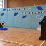 Mężczyzna w garniturze przemawia do mikrofonu przy mównicy na sali gimnastycznej.