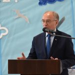 Starszy mężczyzna przemawia do mikrofonu przy mówinicy na sali gimnastycznej.