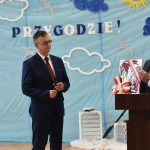 Dwóch mężczyzn stoi na sali gimnastycznej czy mównicy. Jeden z nich trzyma w ręku obraz, który przekazuje na ręce drugiego.