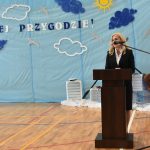 Kobieta o jasnych włosach przemawia do mikrofonu przy mównicy na sali gimnastycznej.