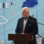 Starszy mężczyzna w siwych włosach przemawia do0 mikrofonu przy mówinicy na sli gimnastycznej.