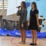 Dwie dziewczyny w sukienkach stoją przed publicznością na sali gimnastycznej. Jedna z nich trzyma mikrofon i mówi wierszyk. Za nimi dekoracja - łódka zrobiona z papieru.