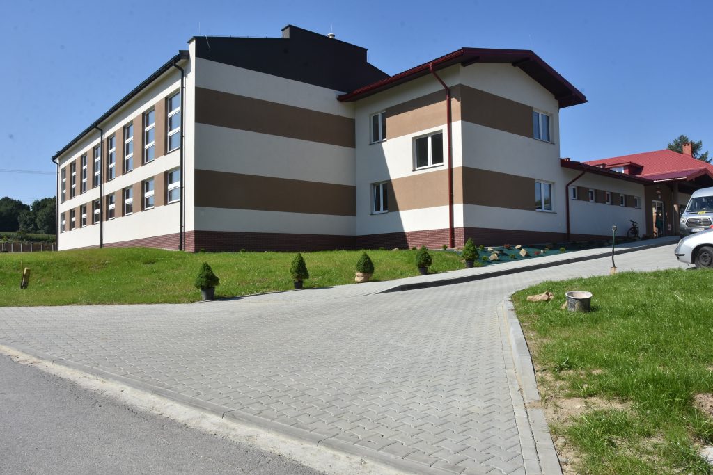 Budynek szkoły w jasnym kolorze elewacji z pasami brązowymi, obok zielony trawnik i wjazd wyłożony kostką.