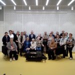 Grupa osób siedzi na krzesłach w jasnym pomieszczeniu.