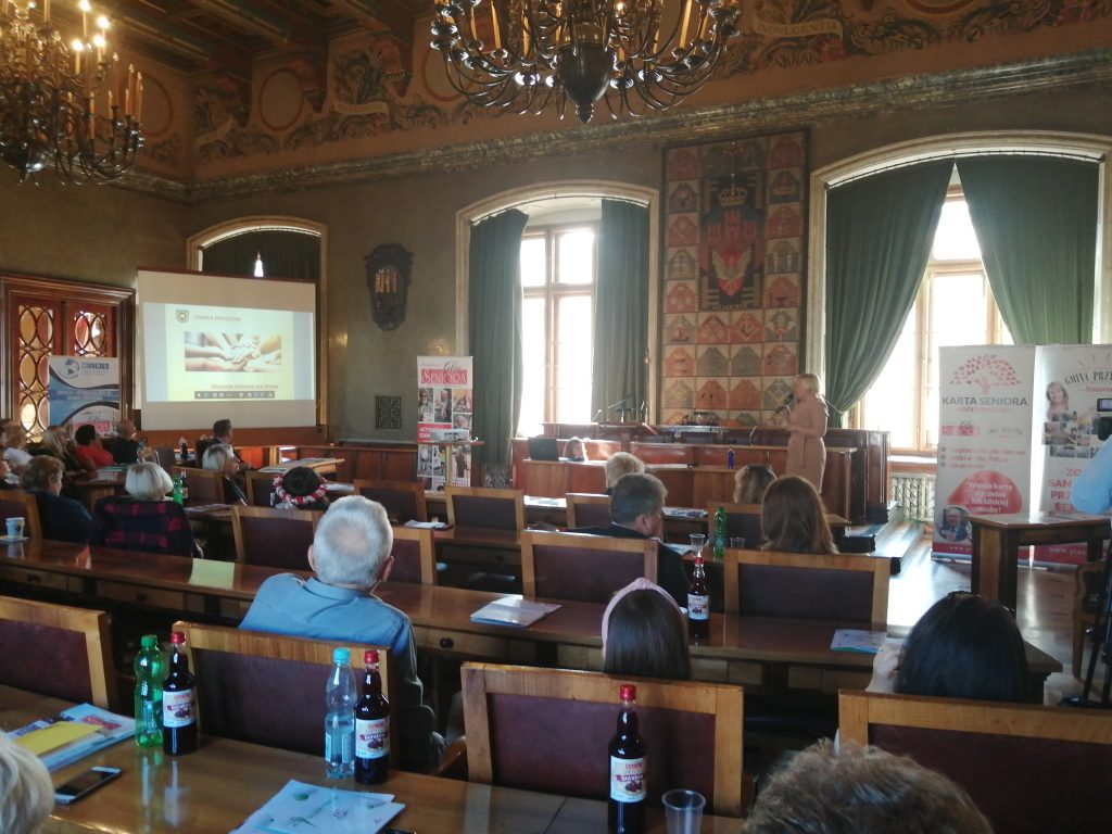 Grupa osób siedzi na dużej sali i przysłuchują się wypowiedziom kobiety, która posługuje się prezentacją multimedialną.