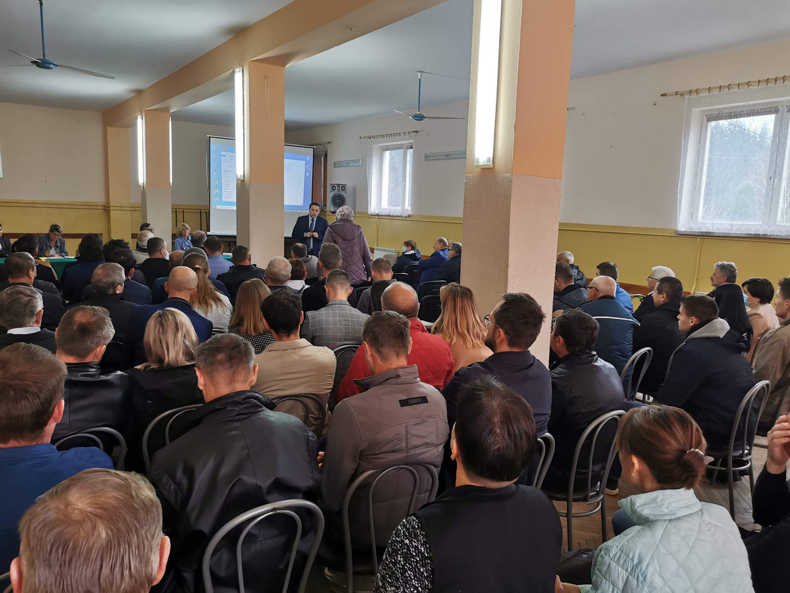 Grupa osób siedzi na zebranie w jasnym pomieszczeniu sali Domu Strażaka, przed nimi stoi mężczyzna który posiłkując się wyświetlaną prezentacją multimedialną przedstawia informacje o stanie gminie.