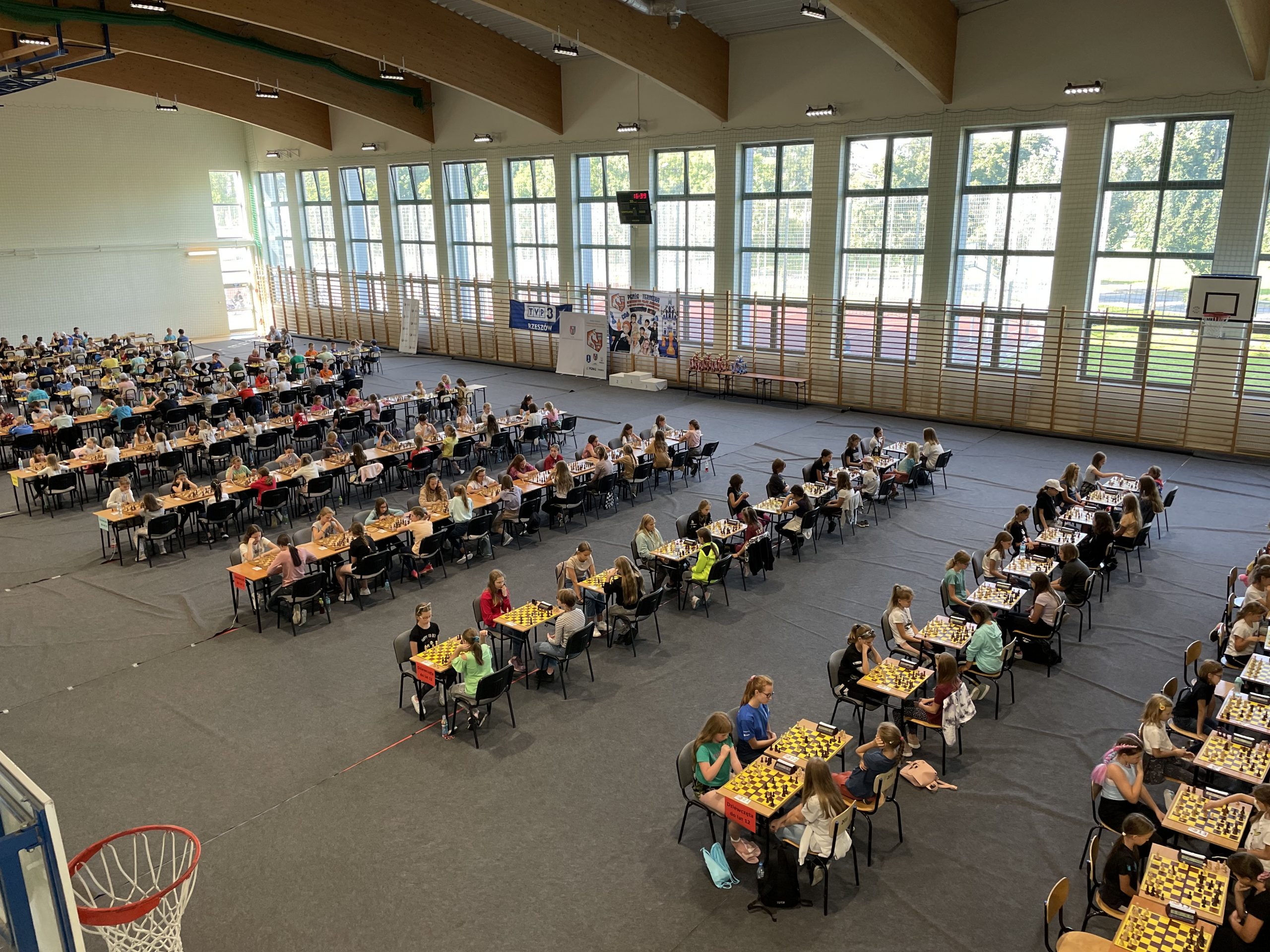 Grupa dzieci siedzą przy stolikach na sali gimnastycznej i grają w szachy,.