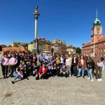 Kilkudziesięciu osobowa grupa młodych ludzi pozuje do zdjęcia podczas wycieczki na rynku miasta. Za nimi ogromne ceglane kamieniczki, mnóstwo przechodniów.