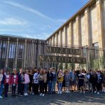 Kilkudziesięciu osobowa grupa młodych ludzi pozuje do zdjęcia podczas wycieczki na tle bramy do jasnego budynku muzeum.