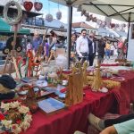 Młody mężczyzna w koszuli z kobietą i wózkiem z dzieckiem stoją na rynku, podczas kiermaszu. Na stołach wyłożonych obrusami czeronymi leżą serwetki z haftami, koszyki słomiane i inne.